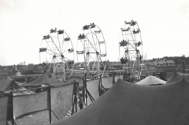 Black and white photo of carnival midway featuring people riding three Ferris wheels set up side by side among many tents and banners