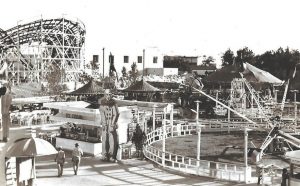 Une photo en noir et blanc d’une fête foraine représentant une montagne russe, un stand de crème glacée et un manège à sensations fortes dont les gondoles montent et descentes tout en tournant