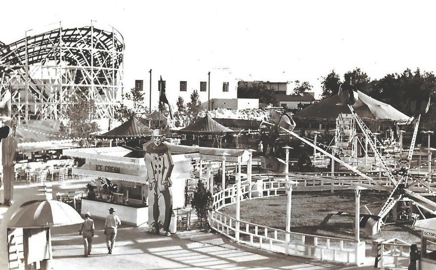 Black and white photo of carnival midway featuring a roller coaster, ice cream stand and up and down spinning thrill ride