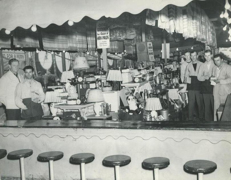 Black and white photo of people standing behind counter for game prizes with a sign that reads One Winner has Choice of Any Prize