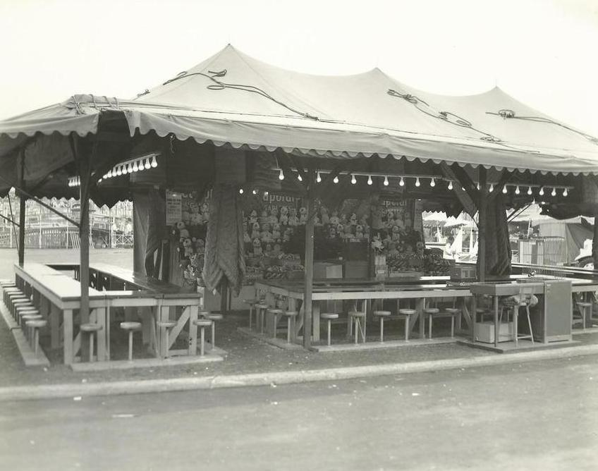 Black and white photo of carnival bingo game set up under a tent with many stools around tables, a wall of stuffed animal prizes and lights all around the top