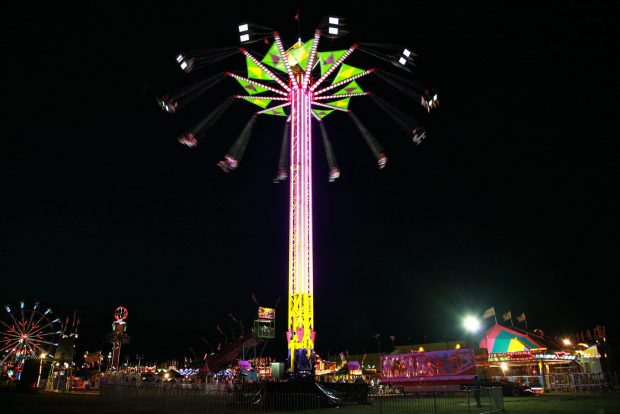 A very tall carnival ride in the middle of a midway, at the top are people swinging around in chairs attached by lines, the ride is lit up under the night sky