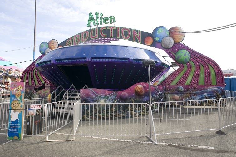Alien Abduction carnival ride, the ride has a round shape with a big doorway to enter inside, there are colourful lights all over the outside and colourful graphics of outer space along the bottom, the WCA lion mascot is on a sign nearby