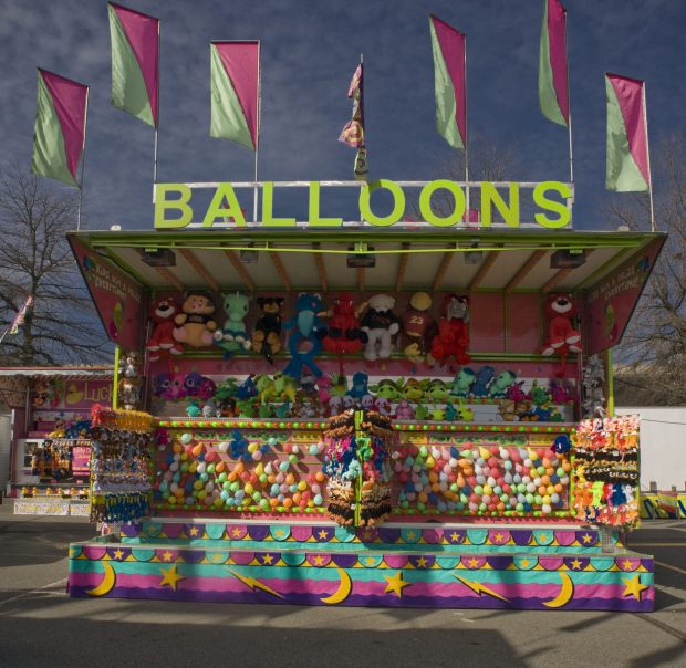 Carnival game concession stand, it is very colourful with flags at the very top, stuffed toys mounted on the wall, balloons for popping and canvas covering the bottom.