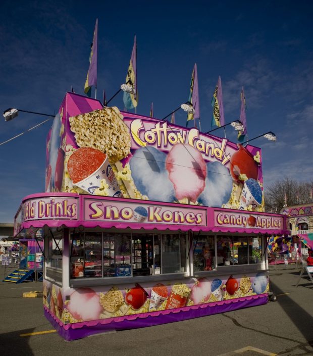 Concession food stand featuring cotton candy, also selling popcorn, candy apples, snow cones and cold drinks. The stand is pink and purple and has colourful graphics of the food all over