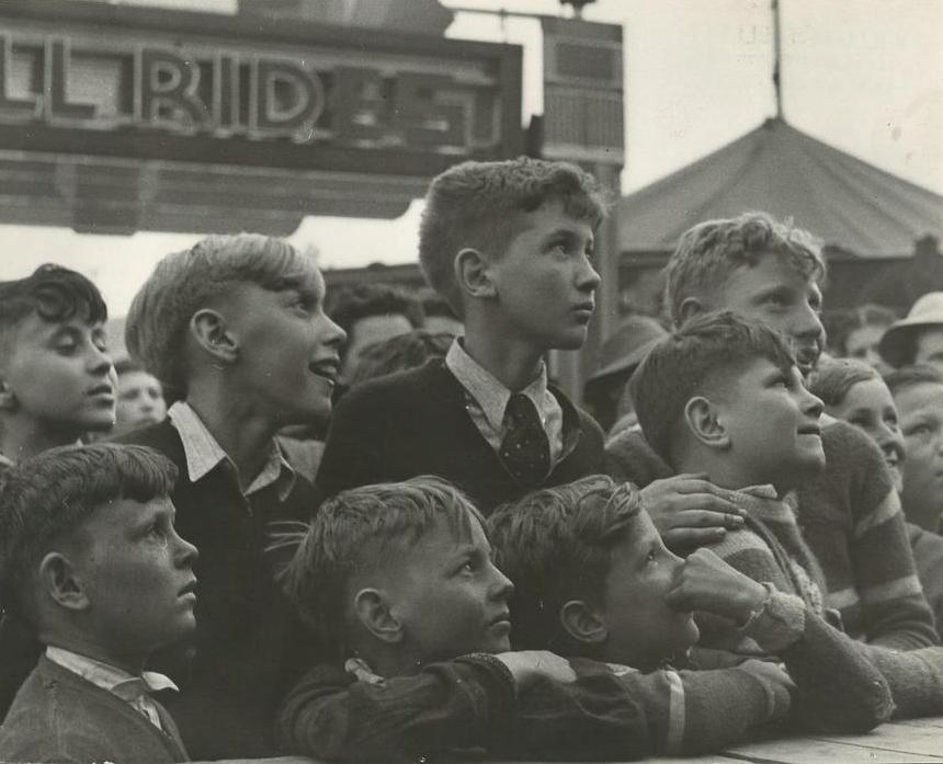 Black and white photo of a close up of young boys gathered together at the carnival, part of sign for thrill rides can be seen in the background