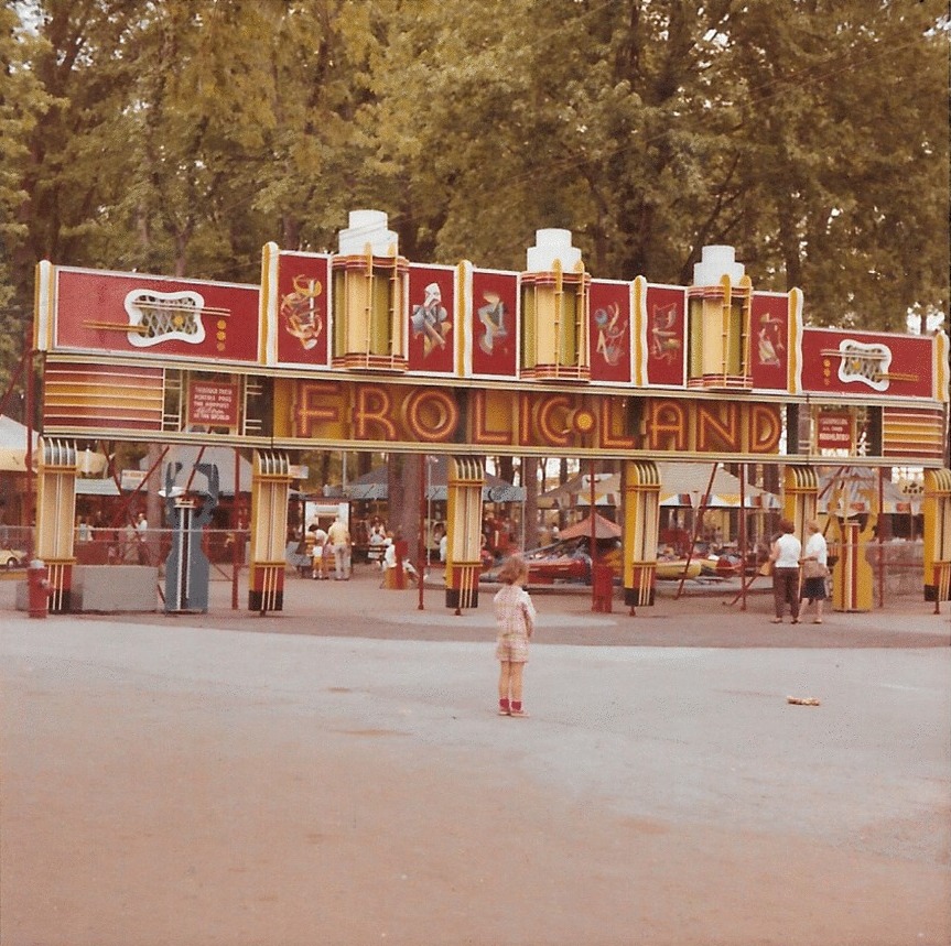 A small young girl stands in front of a large entrance to the carnival midway Frolic Land, there are not many other people around