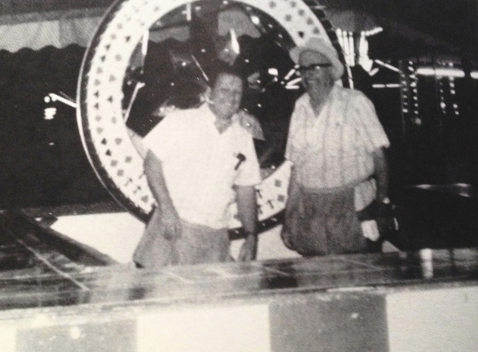 Black and white photo of a middle aged Bingo Hauser smiling and wearing a microphone headset standing beside another man, both behind a carnival game counter and larger spinning wheel is behind them