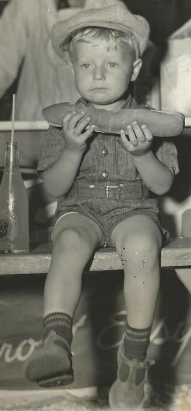 Black and white photo of a small young boy holding a foot-long hot dog, sitting with a bottle of soda beside him