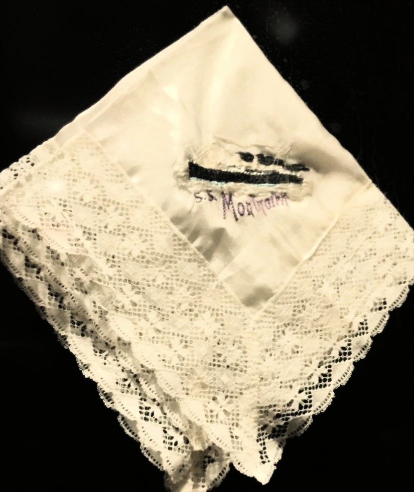 An off-white folded handkerchief with lace edging, there is embroidery in the middle in black and letters in purple