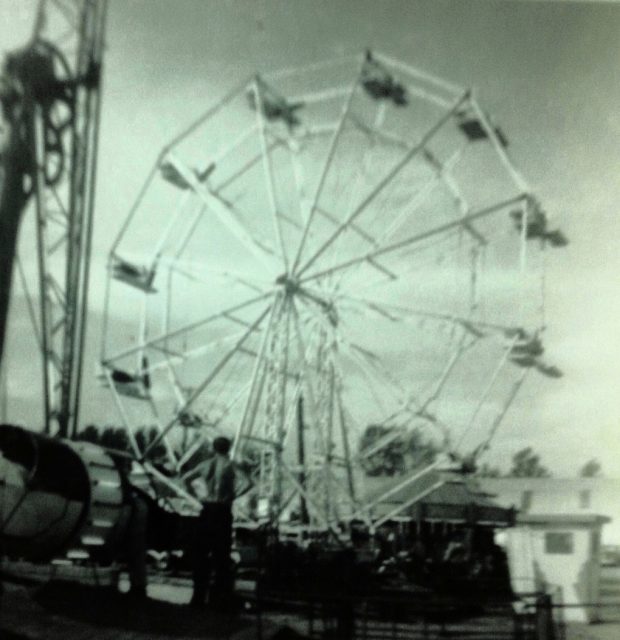 Black and white photo of a Ferris wheel set up at a carnival midway, a shirtless man has his back turned facing the Ferris wheel