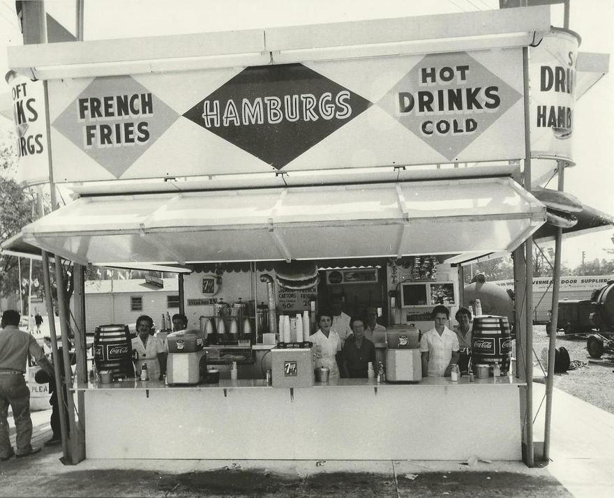 Black and white photo of a food concession stand, the sign says French Fries, Hamburgs and Hot Cold drinks, there are employees behind the counter with some in an all white uniform