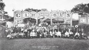 Black and white group photo of many people with a circus, visible is a very obese woman, a band, people in costumes, women in bathing suits and large cut boards with the circus title and one of a painted face with a large open mouth