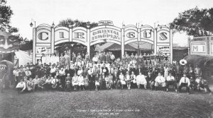 Une photo de groupe en noir et blanc d’un cirque, on peut y voir une femme très obèse, un groupe de musique, des gens en costume, des femmes en maillot de bain, au-dessus du groupe, il y a de grands panneaux sur lesquels le nom du cirque est écrit et un panneau représentant un visage peint avec une bouche ouverte