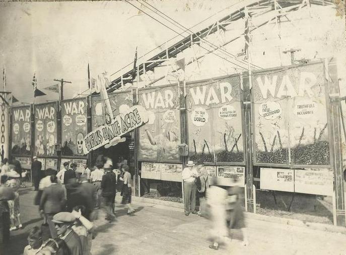 Black and white photo of carnival midway with large painted banners with war written all over, people are crowded around the entrance which has a swinging sign over it that reads Hell's Half Acre