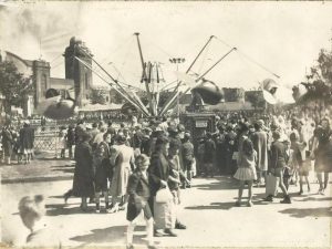 Black and white photo of a large spinning carnival ride at a midway, Flying Scooters can be partially seen