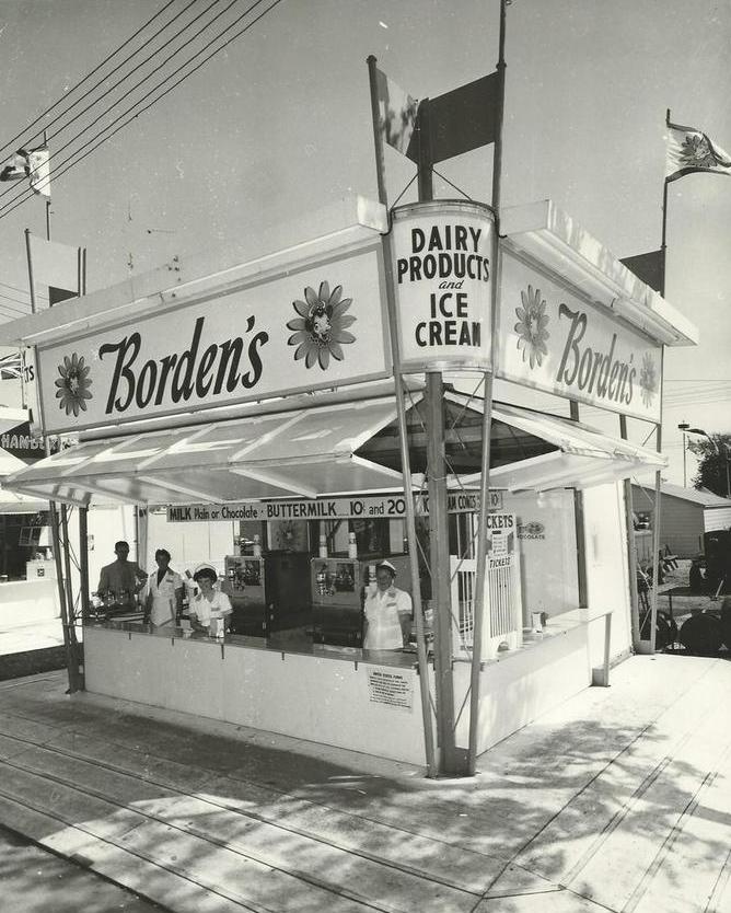 Black and white photo of Borden's ice cream stand, employees are inside with girls in all white uniforms, a sign is visible advertising for butter milk and white or chocolate milk