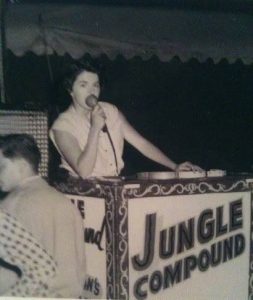 Black and white photo of a woman holding a microphone to her mouth standing behind a podium that says Jungle Compound