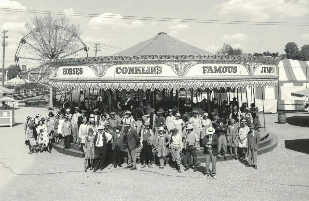 Black and white photo of a group of many people posed in front of a carousel at a midway, a striped tent and Ferris wheel can be seen in the background