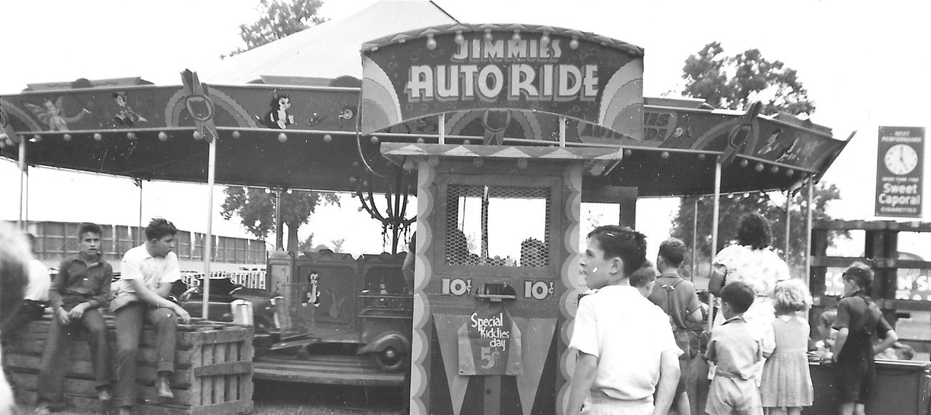 Black and white photo of a few people gathered around a kiddie ride, sign for ride reads Jimmie's Auto Ride, there is also a sign that says Special Kiddies Day five cents