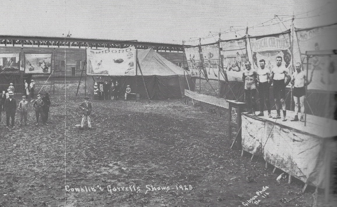 Black and white photo of a carnival bannerline, with people watching shirtless men on a stage, the banner with words physical culture written on it is in the background