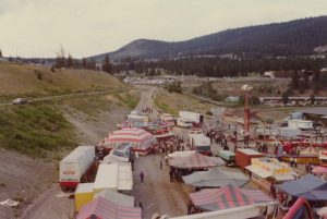 Une fête foraine comprenant plusieurs tentes, manèges et stands, ainsi qu’un camion du WCA, on peut y distinguer le manège Loop-O- Plane et en arrière-plan, une région montagneuse avec beaucoup d’arbres