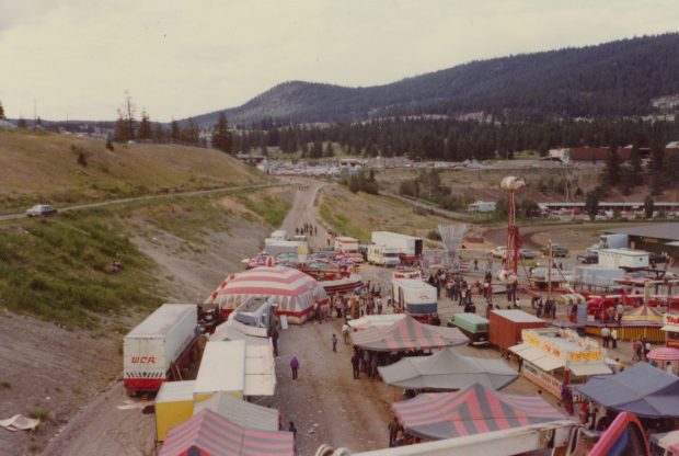 A carnival midway with many tents, rides and concessions stands, a transport truck with WCA on it can be seen, there is also a Loop-O-Plane ride visible, the background is a mountainous area with many trees