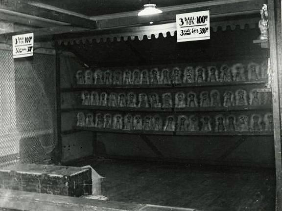 Black and white photo of a carnival cat rack game, a metal rectangular stand with three racks of clowns that can be knocked down, there is a sign that says three balls for one dollar and three cats three dollars