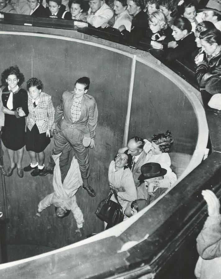 Black and white photo of a large spinning ride, people are stuck to the walls inside the large hallow dome from the acceleration, many people are above looking down to watch