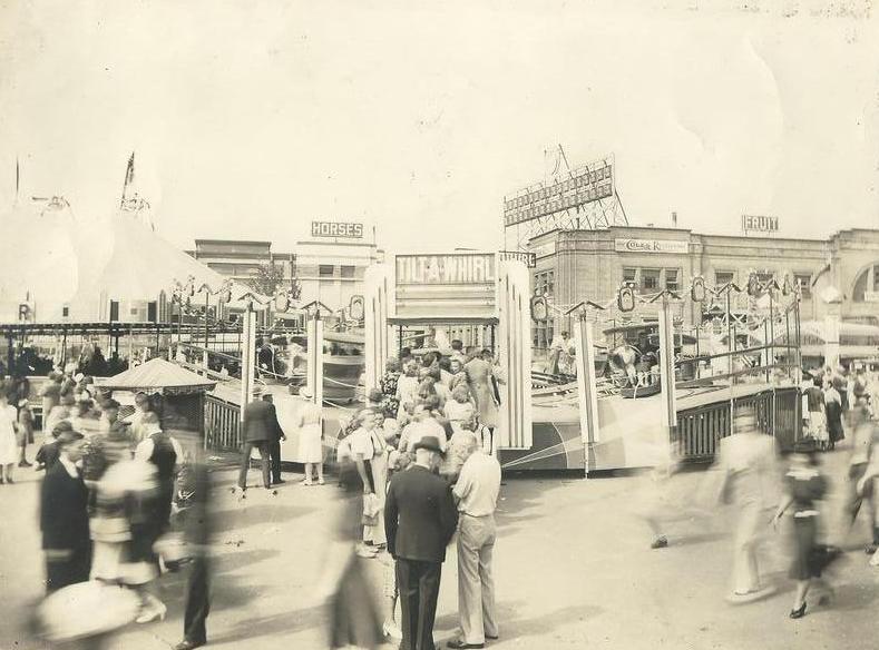 Black and white photo of crowds of people at a carnival midway, featuring people riding a Tilt-a-Whirl ride