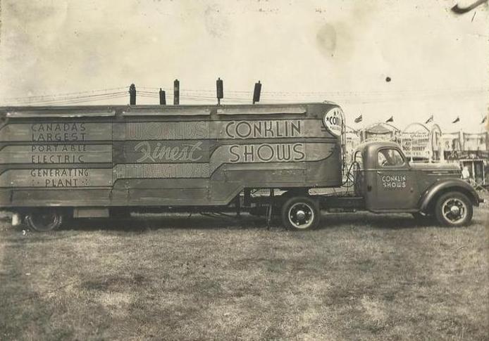 Black and white photo of a large transport vehicle with a portable generator attached, exterior advertises Conklin Shows, carnival banners can be seen in the distance
