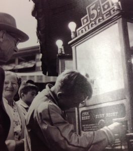 Black and white photo of a young boy playing with a crank five cent digger game, on the machine is says turn right near the controller and coin here where money is put in, people are in the background including an older man behind the boy