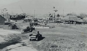 Photo en noir et blanc des attractions d’une fête foraine présentant deux camions à l’avant et plusieurs attractions, y compris une maison du rire (Fun House)