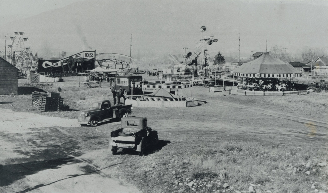 Black and white photo of a carnival midway, two trucks are in front, there is a lot of different areas set up including a Fun House
