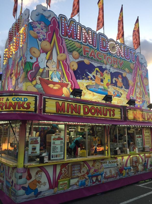 A concession stand for mini donuts, also advertising hot and cold drinks, the stand has colourful graphics and cartoons all over, the sign is lit up