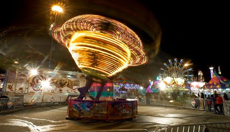 A carnival midway at night, featuring a rapidly spinning lit up ride, a Ferris wheel can be seen in the background