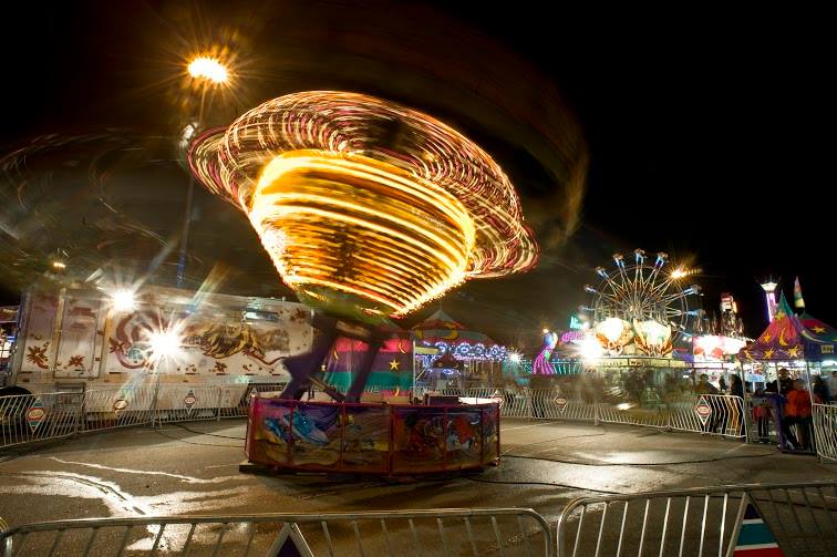 A carnival midway at night, featuring a rapidly spinning lit up ride, a Ferris wheel can be seen in the background