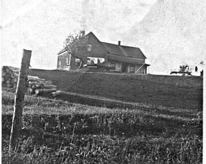 Black and white archival photo of a farm house sitting atop a hill, logs are piles to the left midway down the hill.