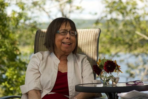 Coloured photograph of Deby Nash Seated with trees and a river behind her.