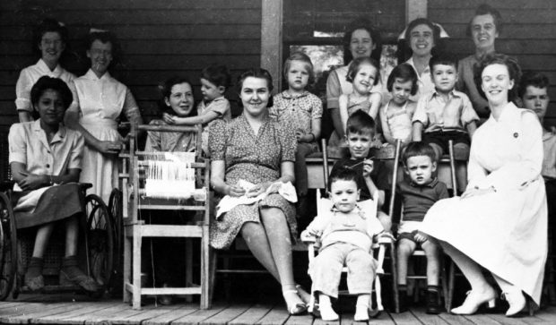 Black and white group photograph of children, nurses, and staff seated on a porch; Edith Henry seated in a wheelchair in front row, first on the left.