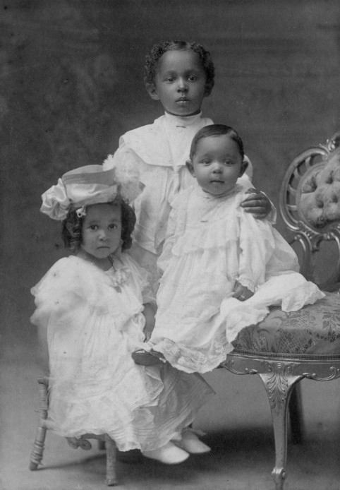 Black and white photo of two young Black girls and one Black boy wearing white.