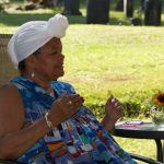 Coloured photograph of Dr. Mary McCarthy-Brandt seated with a green lawn in the background, wearing a blue dress and white head wrap.