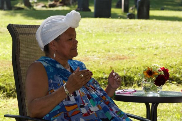 Coloured photograph of Dr. Mary McCarthy-Brandt seated with a green lawn in the background, wearing a blue dress and white head wrap.