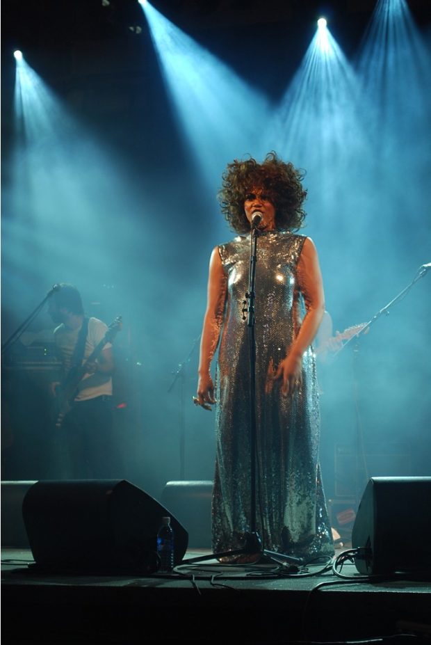 Recent photo of a woman standing and singing on a brightly lit stage wearing a full length dress. A bass player is in the background.
