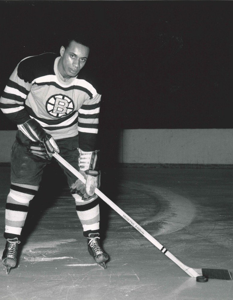 Black and white photo of Willie O'Ree on the ice in his hockey gear, wearing Boston Bruins uniform, holding a hockey stick.