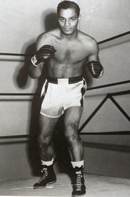 Black and white photograph of a Black man in a boxing pose wearing shorts and boxing gloves.