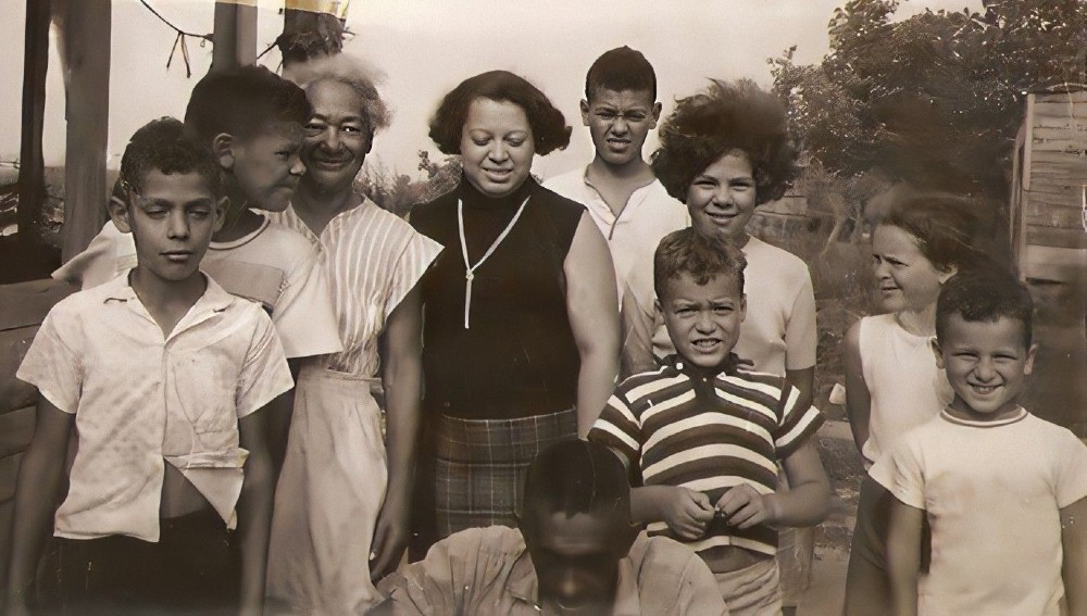 Black and white archival photo of a family of 10 including adults and children standing in front of a porch.