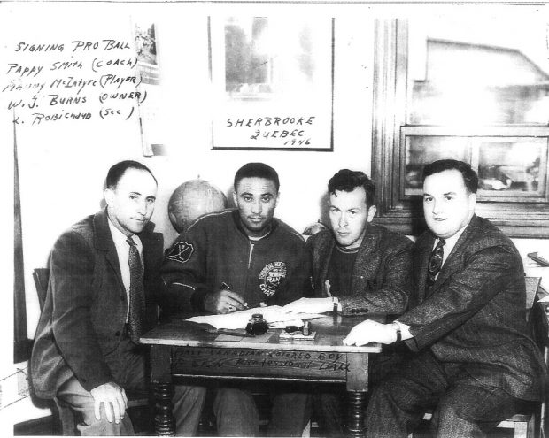 Black and white photo of four men sitting at a table, Manny McIntyre second from the left is shown signing a pro ball contract.