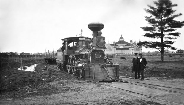 Black and white archival photo showing a railway engine equipped with the Temple & Miller flange, two men wearing suits and top hats stand to the right.