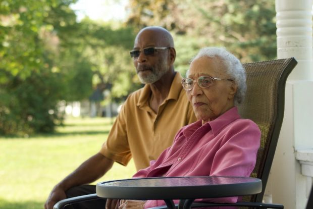 A colour photo of an elderly Black woman wearing a pink shirt seated, behind her is a Black man with a yellow shirt.